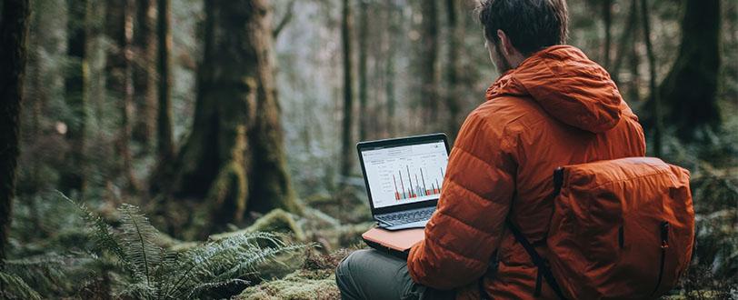 En person med orange jacka och ryggsäck sitter i skogen och arbetar vid en bärbar dator som visar diagram.